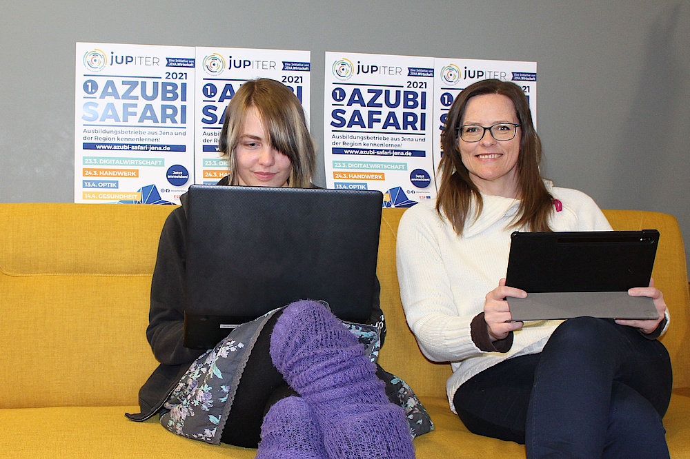 Zwei Frauen sitzen auf gelber Couch und haben einen Laptop und ein Tablet in der Hand. Die linke Frau guckt auf das Endgerät und die rechte Frau in die Kamera. 