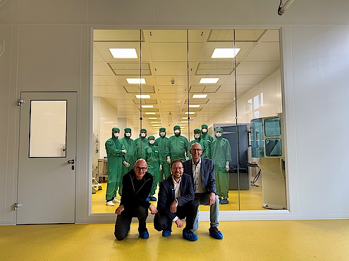 Drei Männer im Vordergrund, dahinter Glaswand und 8 Menschen in grünen Schutzanzügen mit Kapuze und weißem Mundschutz