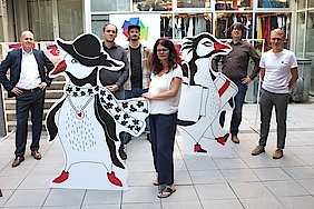 Gruppe von sechs Personen stehen versetzt, dazwischen mannshohe Pinguin Pappfiguren