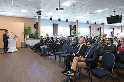Vortragsraum, zahlreiche Menschen sitzen auf Stühlen nebeneinander und schauen auf Redner links im Bild
