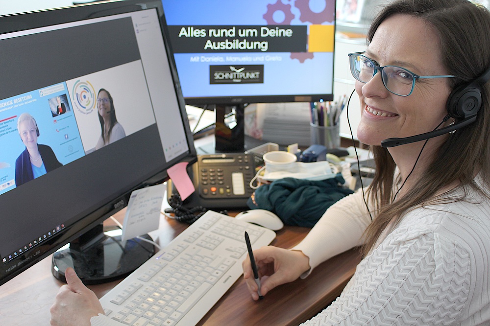 Frau mit braunen Haaren, rechts im Bild, blickt in Kamera und lächelt, links im Bild sind 2 Bildschirme