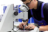 Ein junger Mann in Blaumann arbeitet unter einem Mikroskop an einer Optik.