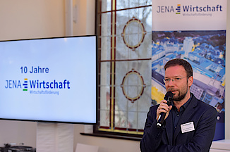 Eine Person spricht in ein Mikrophon, im Hintergrund ein Monitor mit dem JenaWirtschaft-Logo.